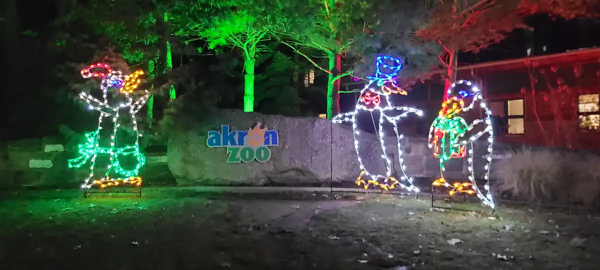 A nighttime photo of the Akron Zoo entrance, featuring a large stone sign with the zoo's name and logo. Three illuminated…