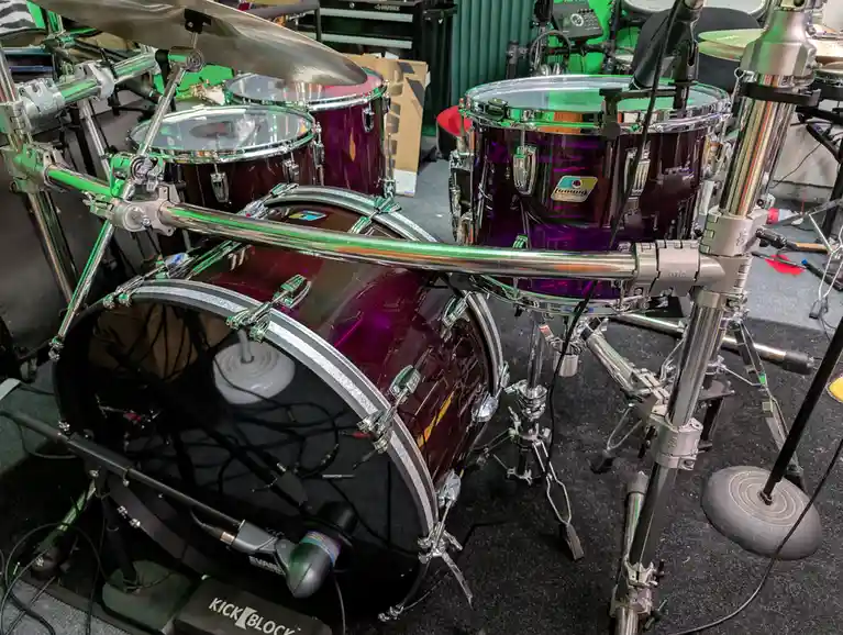 A close-up photo of a shiny, purple-finished drum set with chrome hardware, featuring a bass drum, toms, and cymbals…