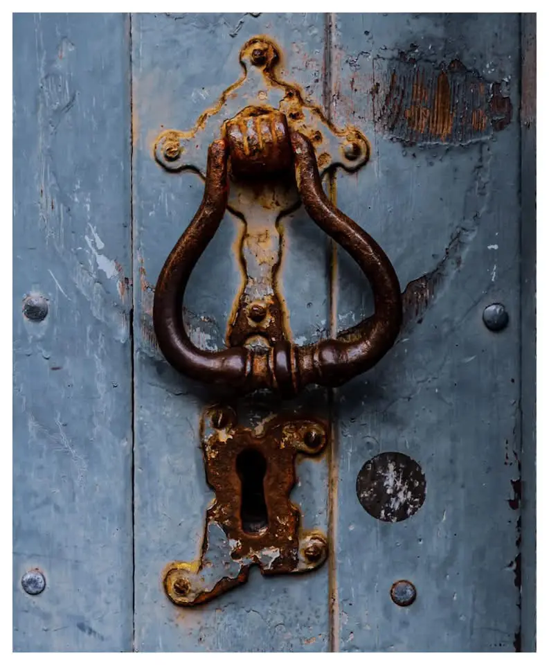 A close-up of a heavily rusted, ornate metal door knocker and keyhole on a weathered blue wooden door, with peeling paint…