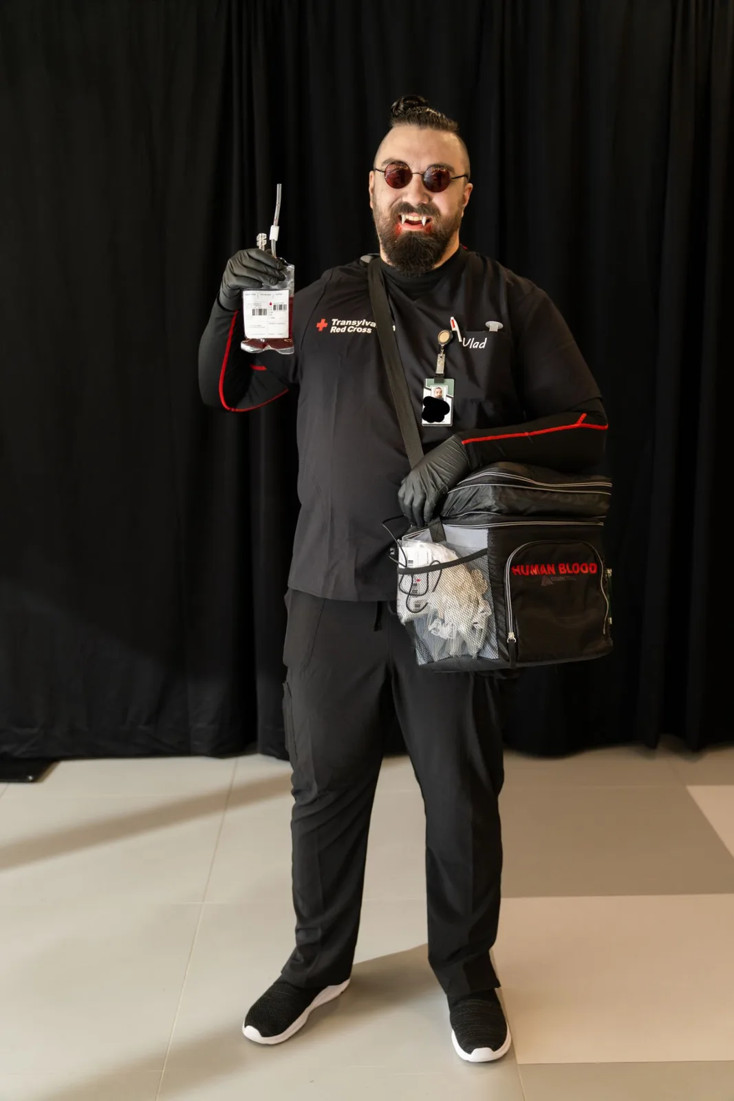 A man dressed as a vampire in black scrubs with 'Transylvania Red Cross' on the chest, holding a blood bag and carrying a…