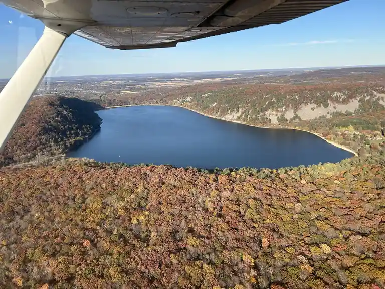 An aerial photo taken from inside a small airplane shows a scenic view of a lake surrounded by forests in autumn colors. The…