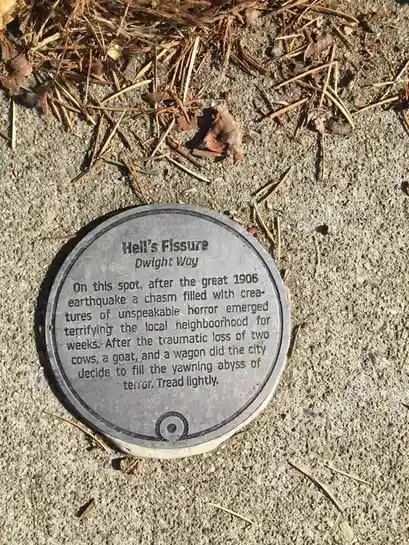 A circular metal plaque embedded in concrete pavement, surrounded by dry pine needles and leaves, reads 'Hell's Fissure…