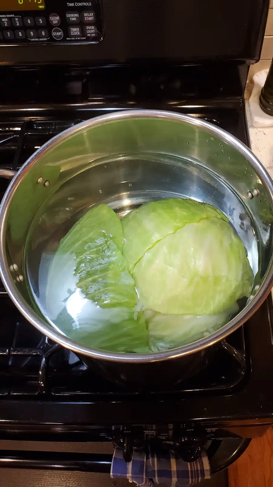 A whole green cabbage is submerged in water inside a large stainless steel pot sitting on a black stovetop. The pot is on a…