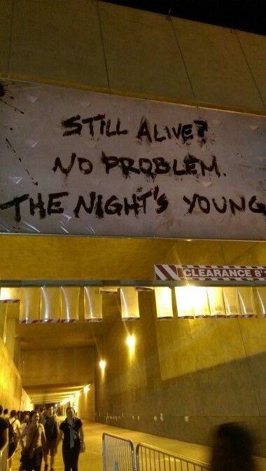 A nighttime photo shows a large white banner with handwritten black text reading 'STILL ALIVE? NO PROBLEM. THE NIGHT'S…
