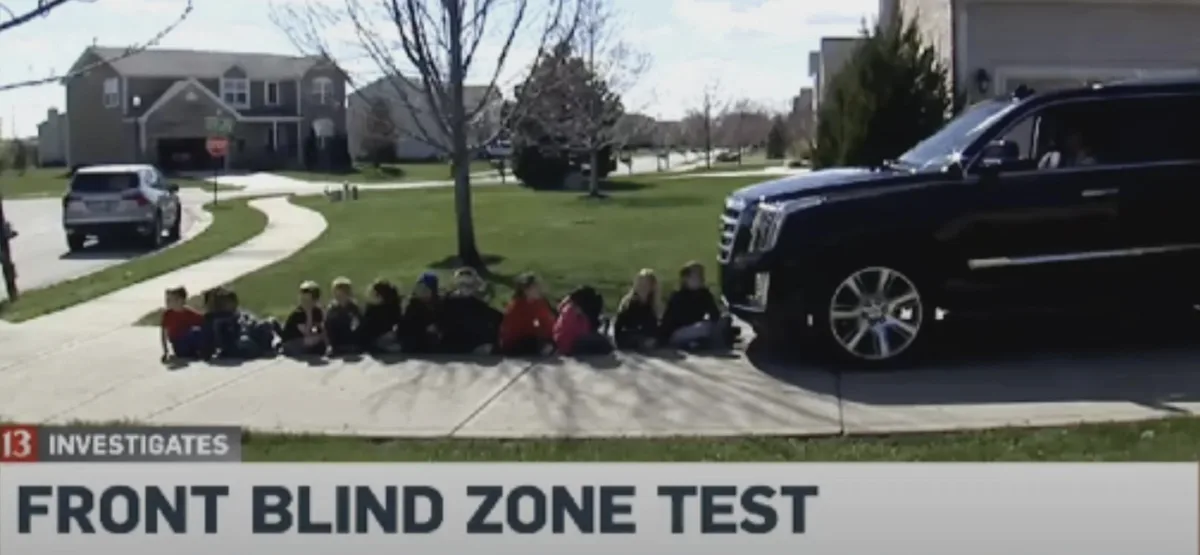 A line of children sits on a sidewalk next to a large black SUV, with a news banner reading 'FRONT BLIND ZONE TEST' at the…