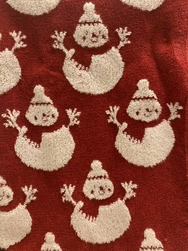 A close-up of a red, textured fabric, likely a towel or rug, featuring a repeating pattern of white, fluffy snowmen. Each…