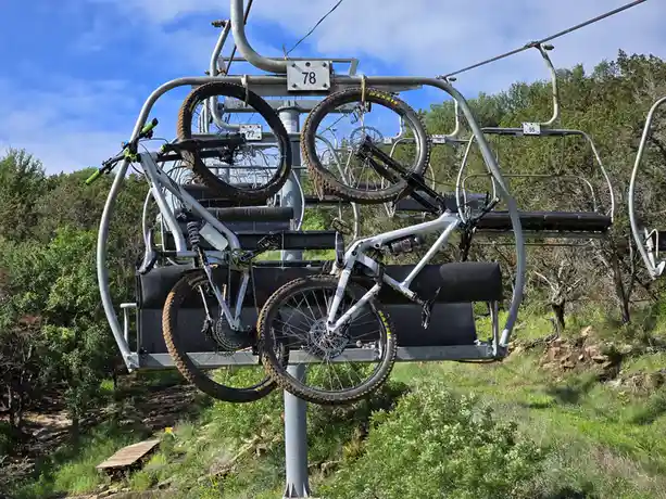 Two mountain bikes are strapped onto a ski lift chair, suspended high above a forested hillside under a blue sky. The bikes…