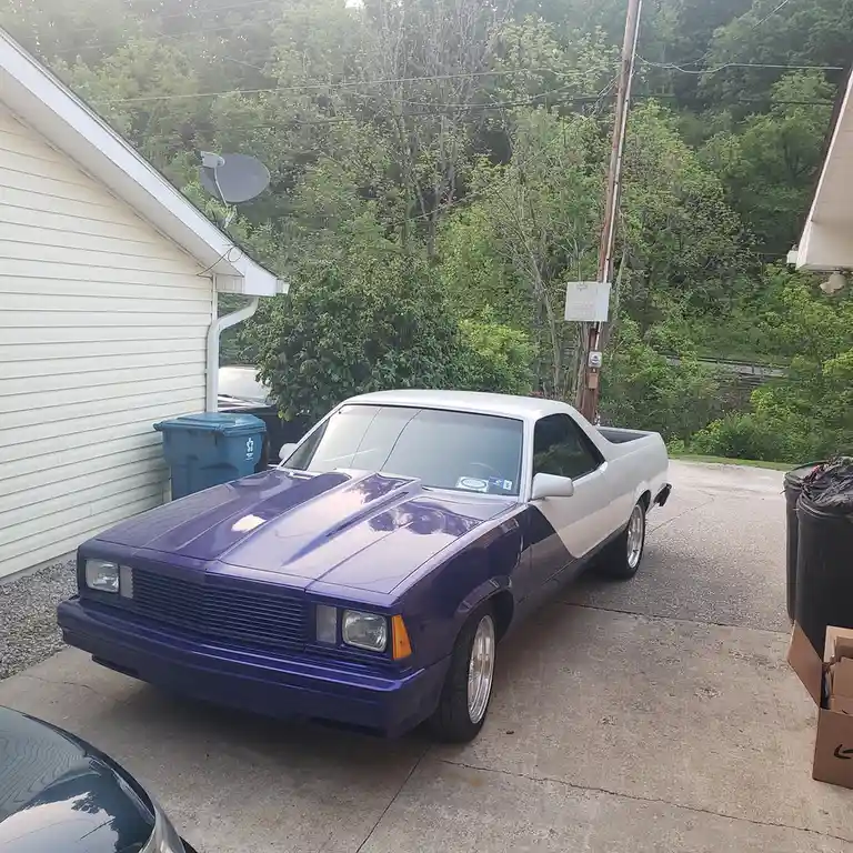 A two-tone purple and white classic Chevrolet El Camino is parked in a residential driveway next to a house with siding and…