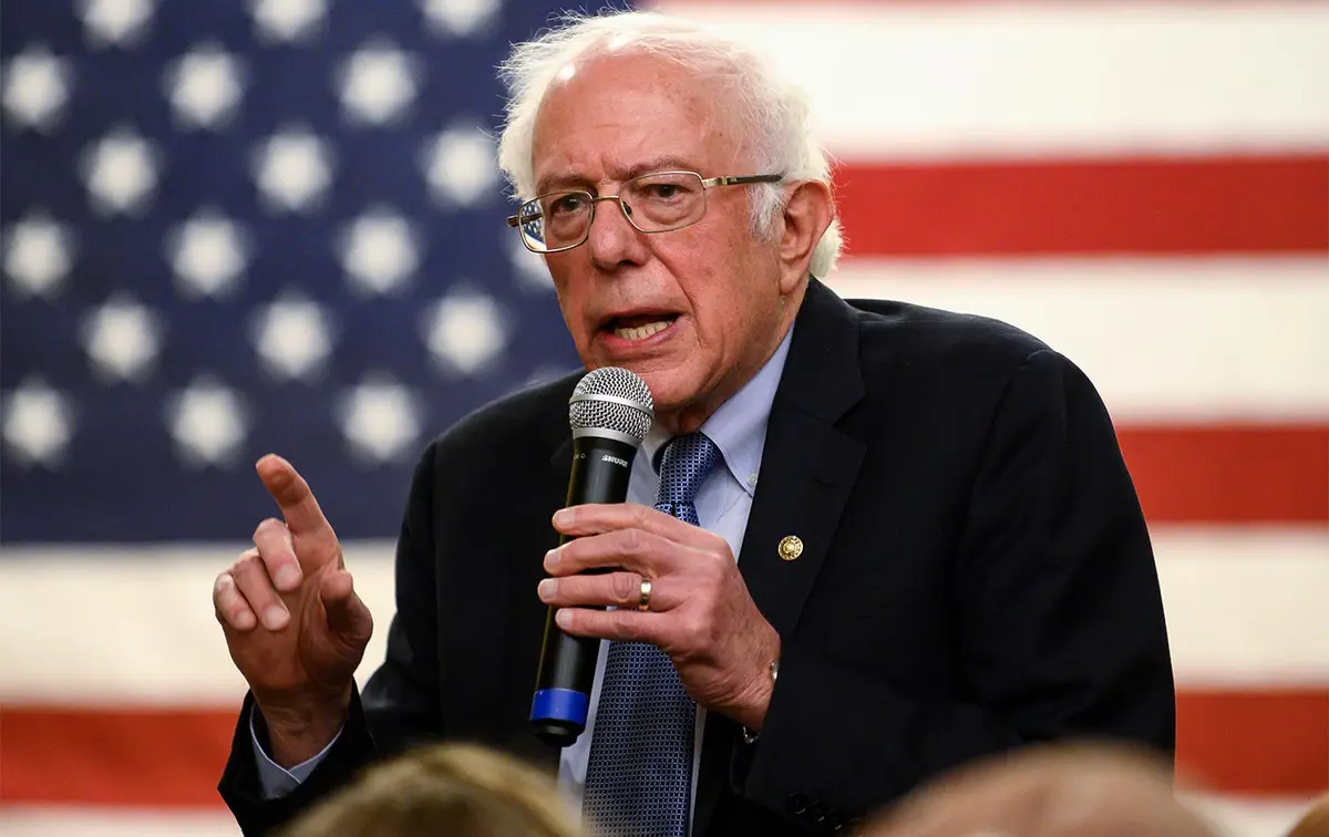 a photo of bernie sanders speaking into a microphone while gesturing with his left hand, standing in front of an american…