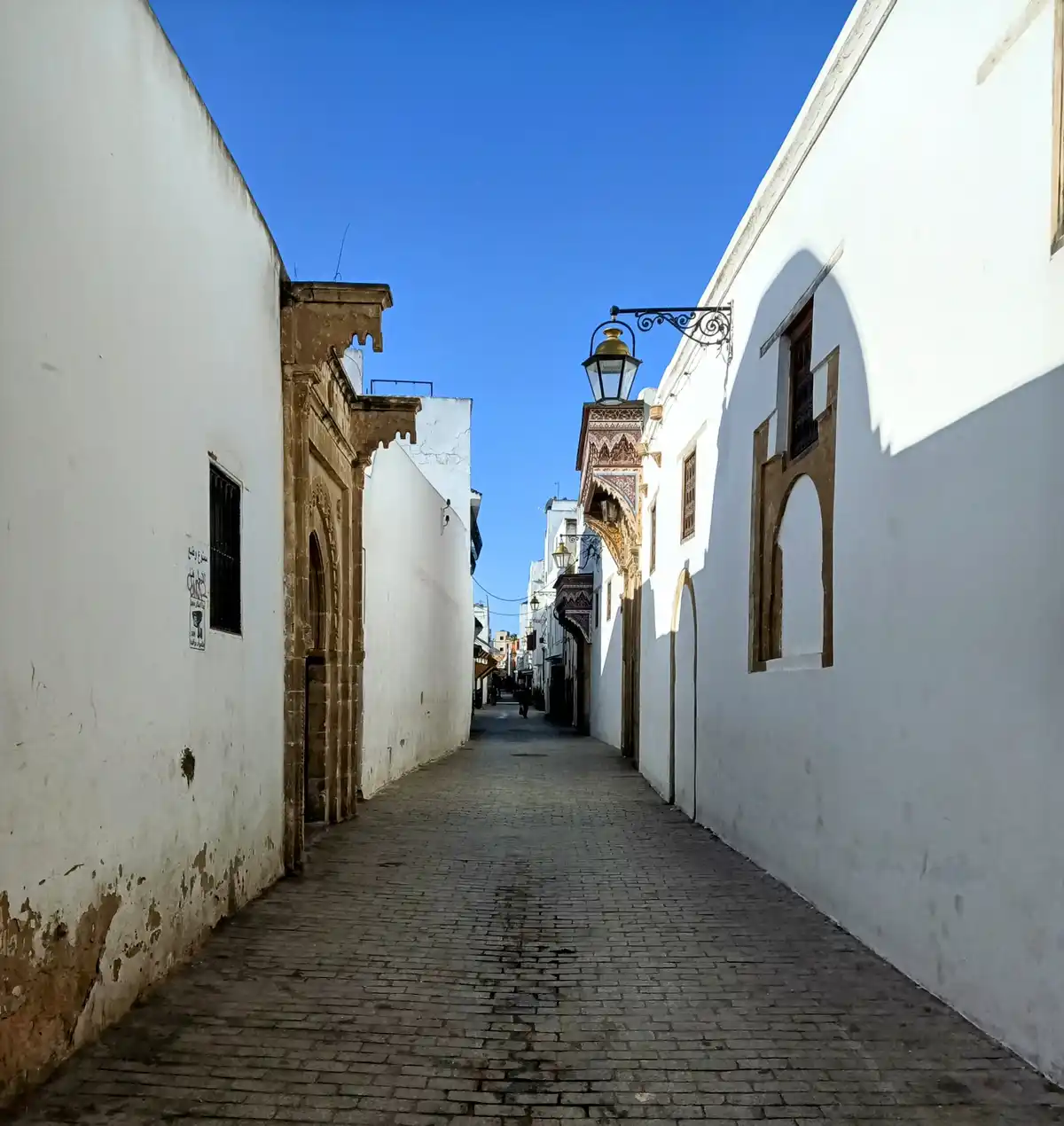 A narrow, sunlit alleyway in Morocco with white-washed walls, ornate arched doorways, and traditional lanterns, overlaid…