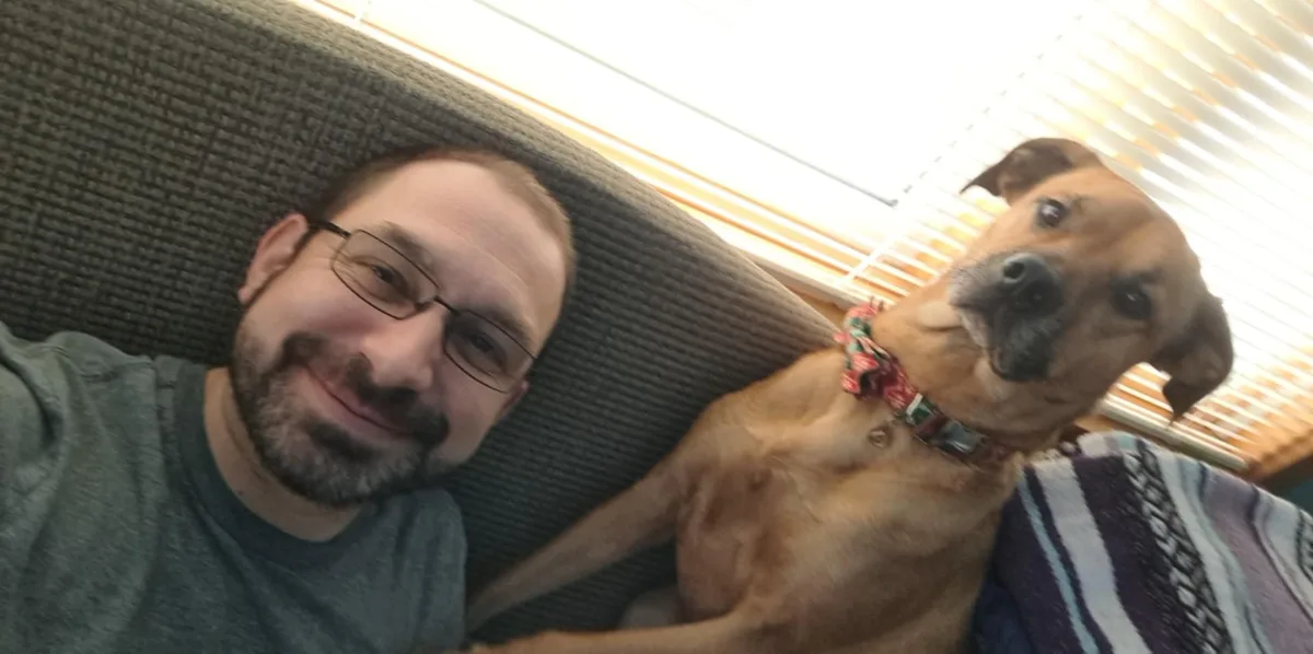 A man with glasses and a beard smiles while lying on a couch next to a brown dog wearing a patterned collar. The dog looks…