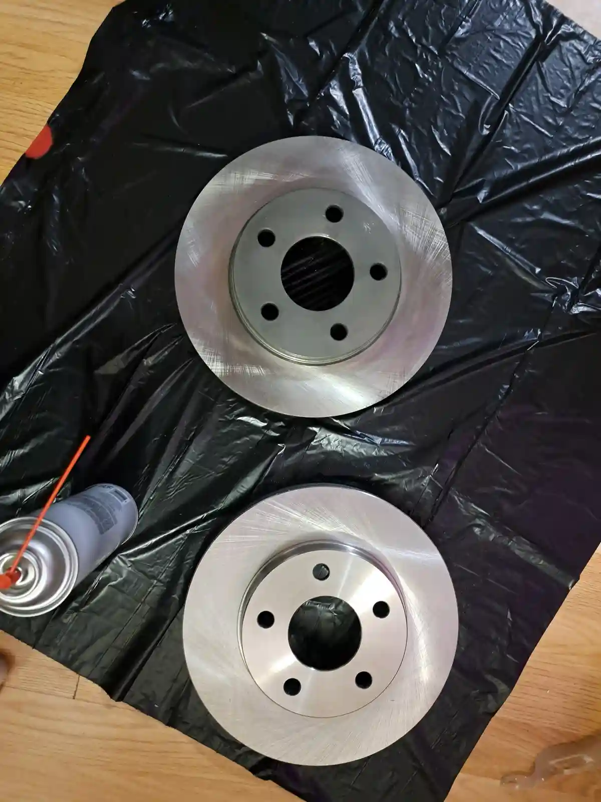 Two shiny, freshly painted brake rotors sit on a black plastic sheet on a wooden floor, next to a can of spray paint with an…