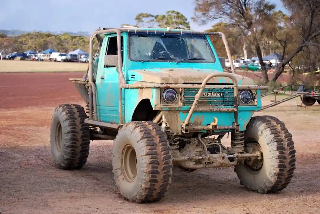 A heavily modified, mud-splattered turquoise Suzuki off-road truck with oversized tires and a protective roll cage stands on…