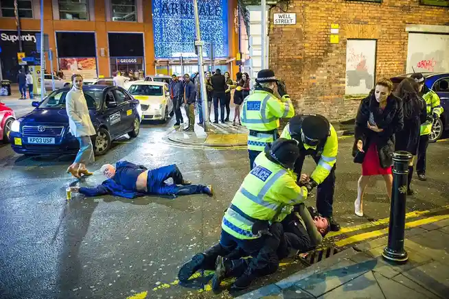 A nighttime street scene in a city with police officers arresting a man on the pavement while another man in a blue jacket…