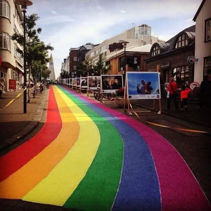A wide, paved city street is painted with a vibrant, curving rainbow stretching from the foreground toward the horizon…
