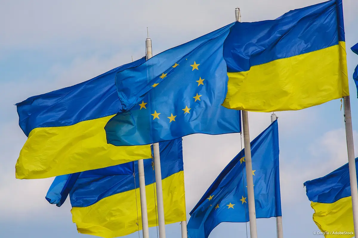 Multiple Ukrainian flags and European Union flags are waving together on poles against a cloudy sky, with the EU flag…