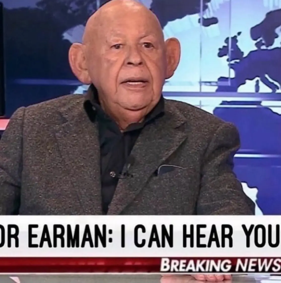 An older bald man with large ears is shown on a news set with a world map background. A news ticker at the bottom reads…