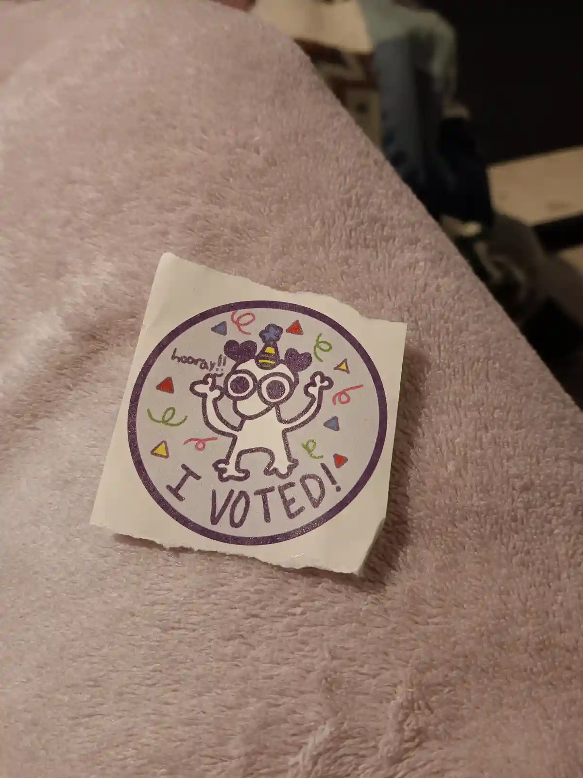 A hand-drawn, circular sticker with a purple border sits on a fluffy pink blanket. The sticker features a cartoon character…