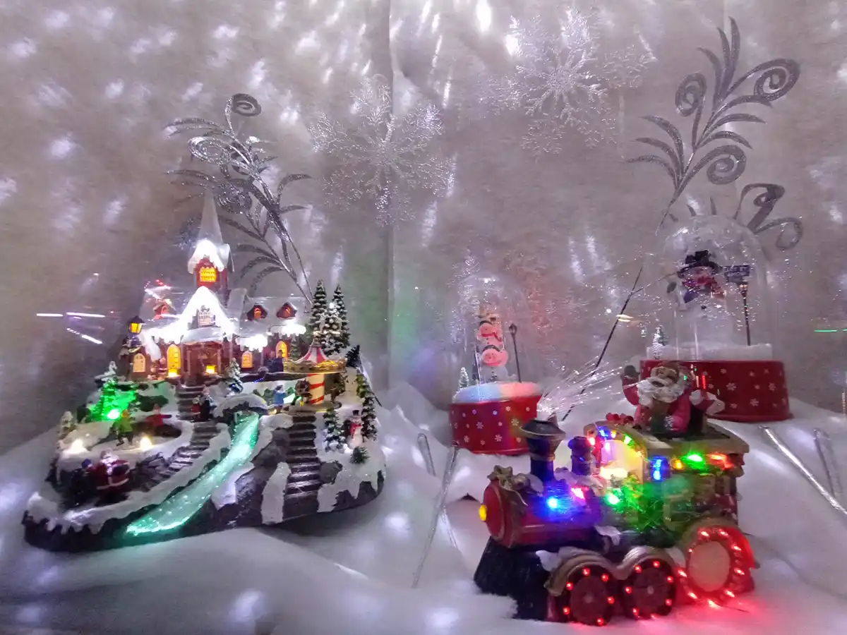 A festive holiday display features a glowing Christmas village diorama with snow-covered buildings and trees, next to a…