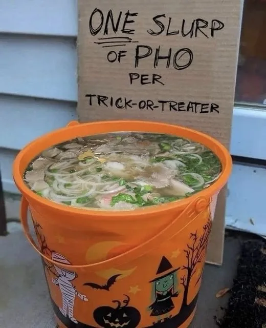 An orange Halloween bucket, decorated with pumpkins, bats, and a witch, is filled with pho soup. Behind it, a cardboard sign…