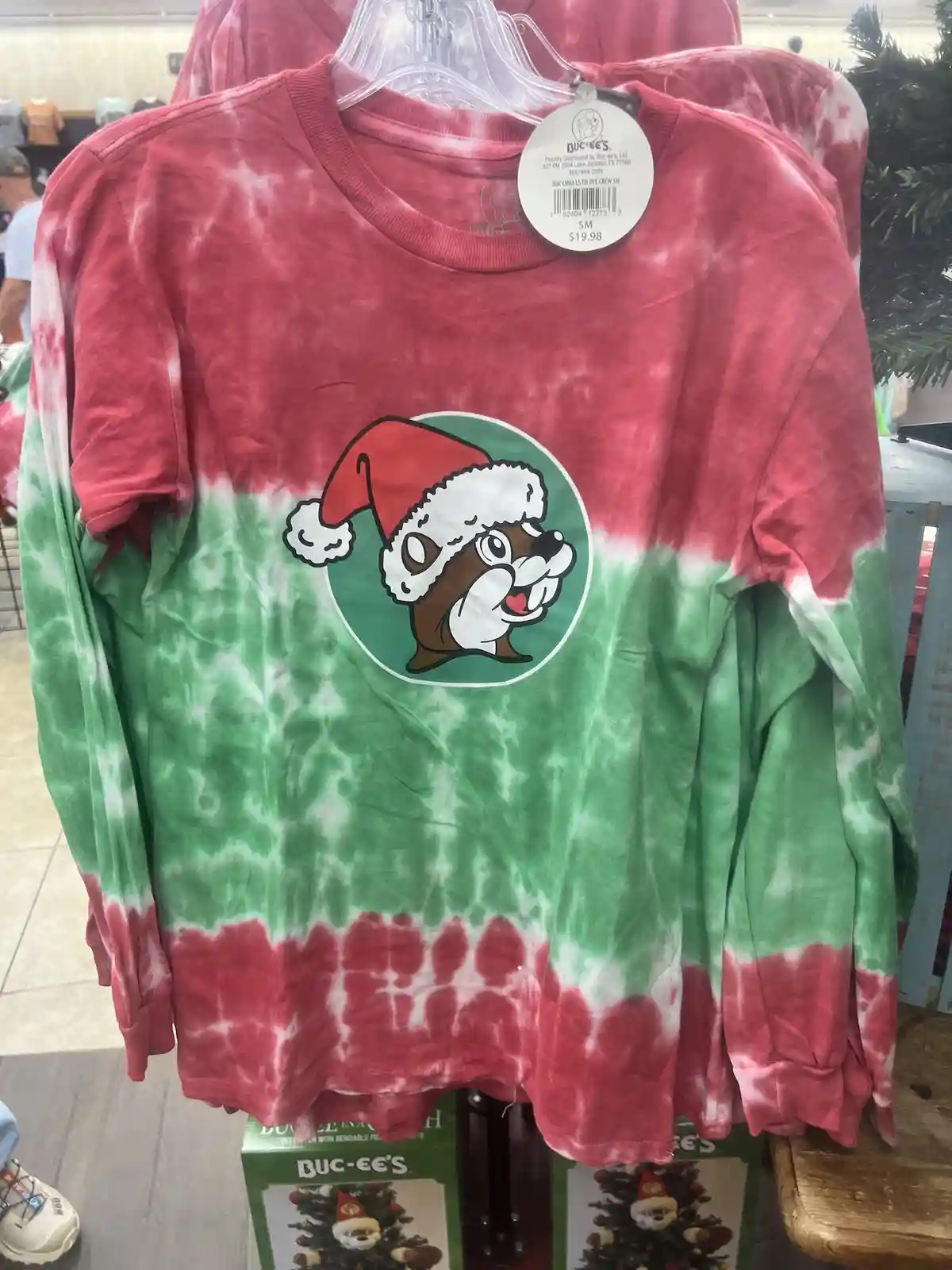 A red and green tie-dye long-sleeve shirt hangs on a hanger in a store, featuring a circular graphic of a cartoon beaver…