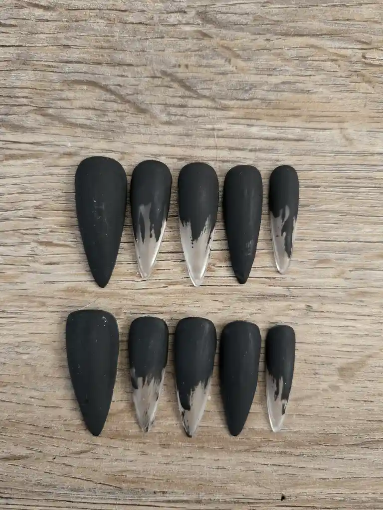 Ten long, stiletto-style false nails arranged in two rows on a wooden surface; five are matte black, and five have a…
