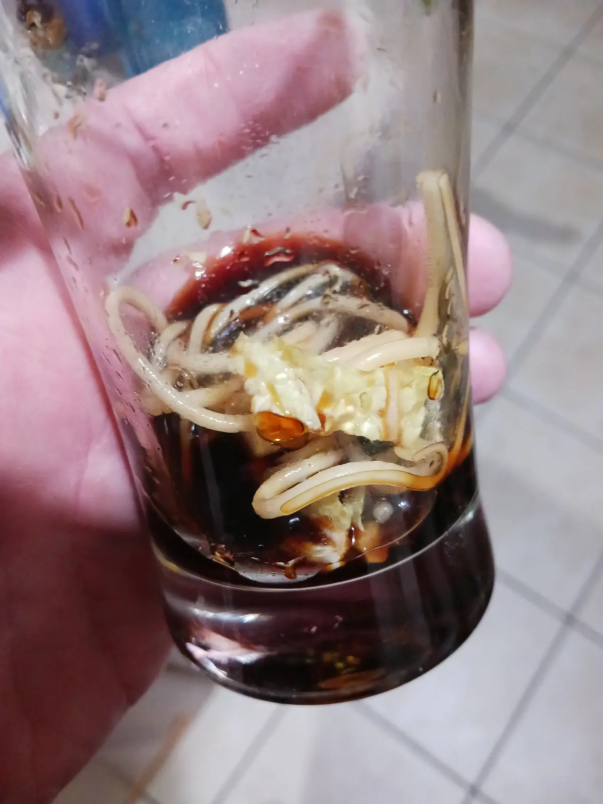 A hand holds a small, clear shot glass containing dark broth, noodles, and a piece of cooked egg, resembling a miniature…