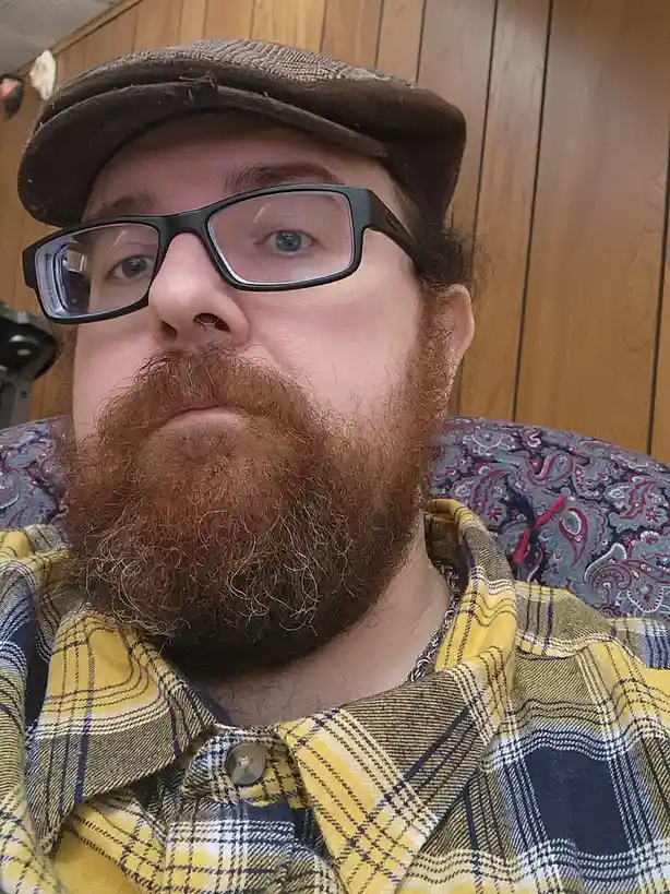 A close-up selfie of a man with a thick, reddish-brown beard, wearing black-rimmed glasses and a brown flat cap, looking…
