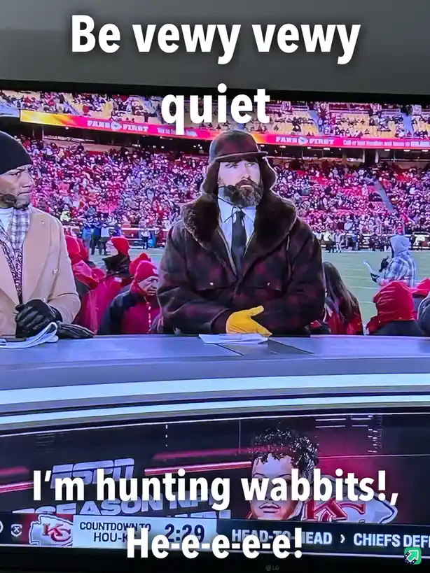 A screenshot of a sports broadcast shows two commentators at a desk, one wearing a fur-lined coat and hat, looking intensely…