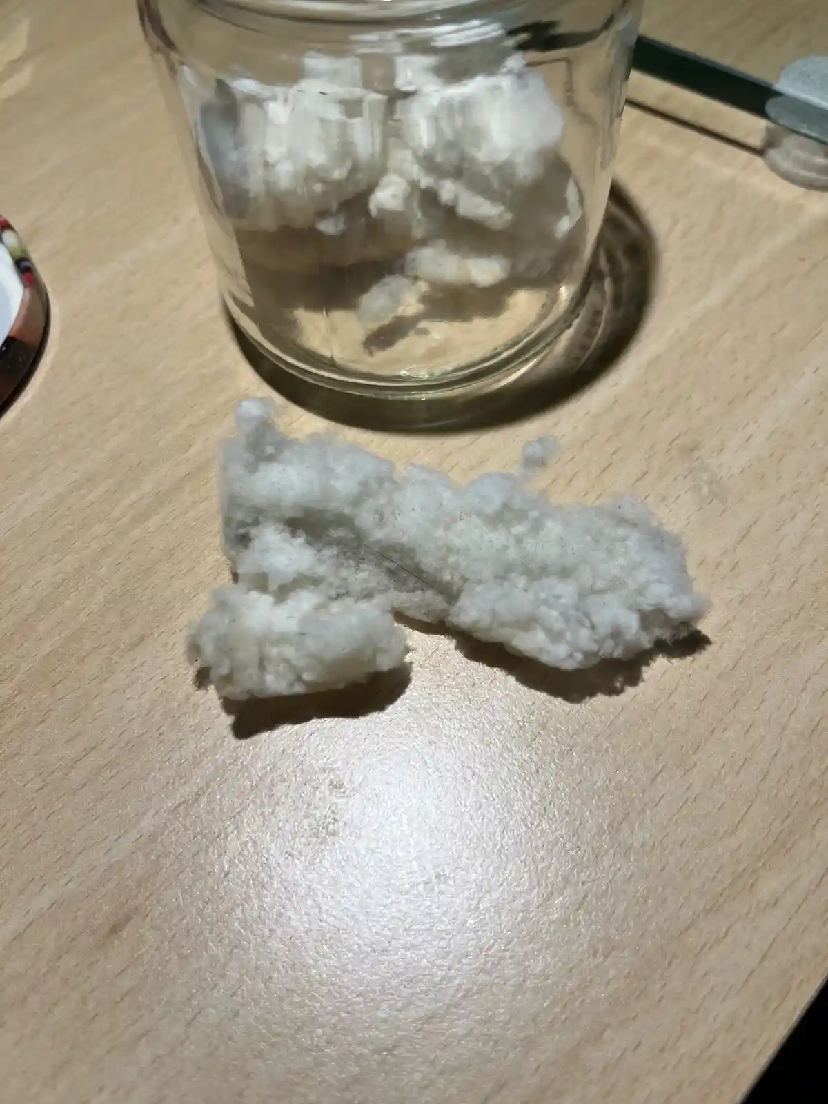 A close-up photo shows a white, fluffy cotton ball resting on a light wooden table, with a clear glass jar containing more…