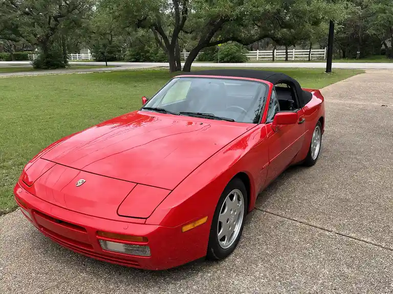 A red Porsche 944 convertible with a black soft top is parked on a gravel driveway next to green grass and trees. The car's…