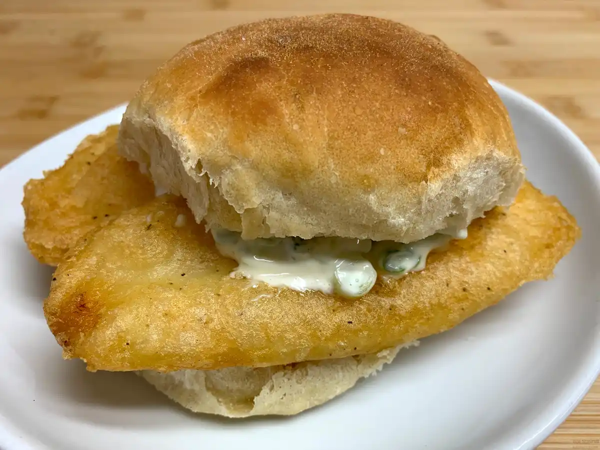 A close-up photo of a golden-brown fried fish fillet sandwiched between two halves of a soft, round bread roll, with a…