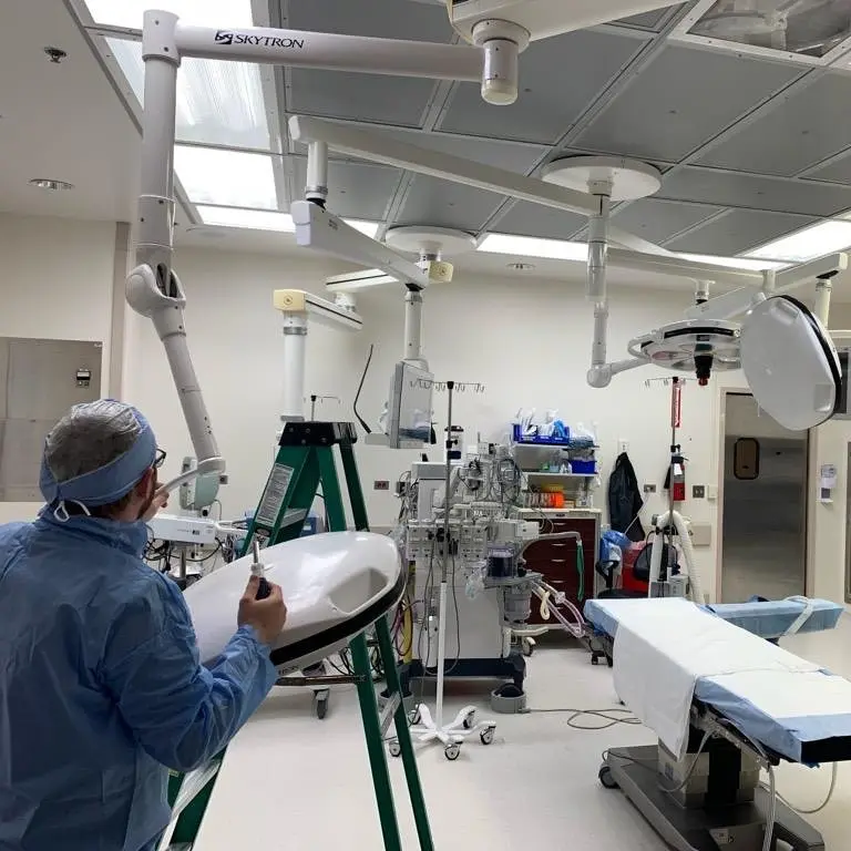A person in blue surgical scrubs and a surgical cap is standing in an operating room, holding a surgical instrument and…