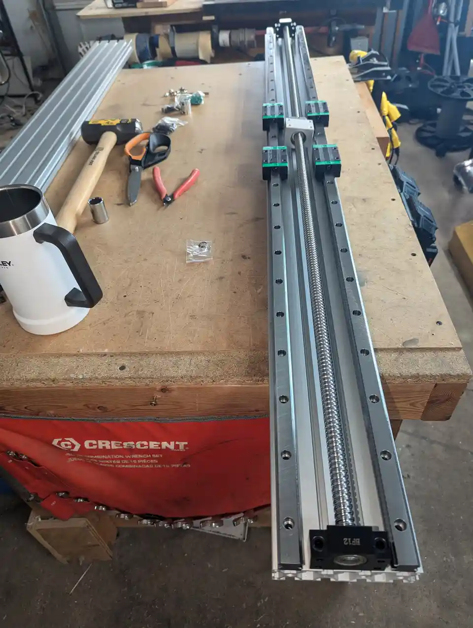 A workshop workbench holds a long, assembled linear rail system with a threaded rod and carriage assemblies, next to a white…
