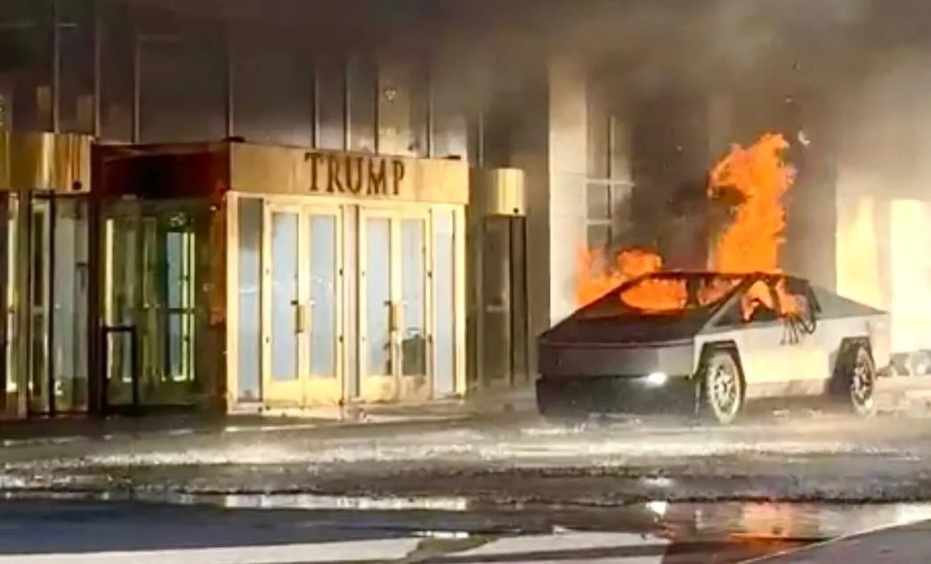 A black Tesla Cybertruck is engulfed in flames on a wet street outside a building with a prominent 'TRUMP' sign above its…