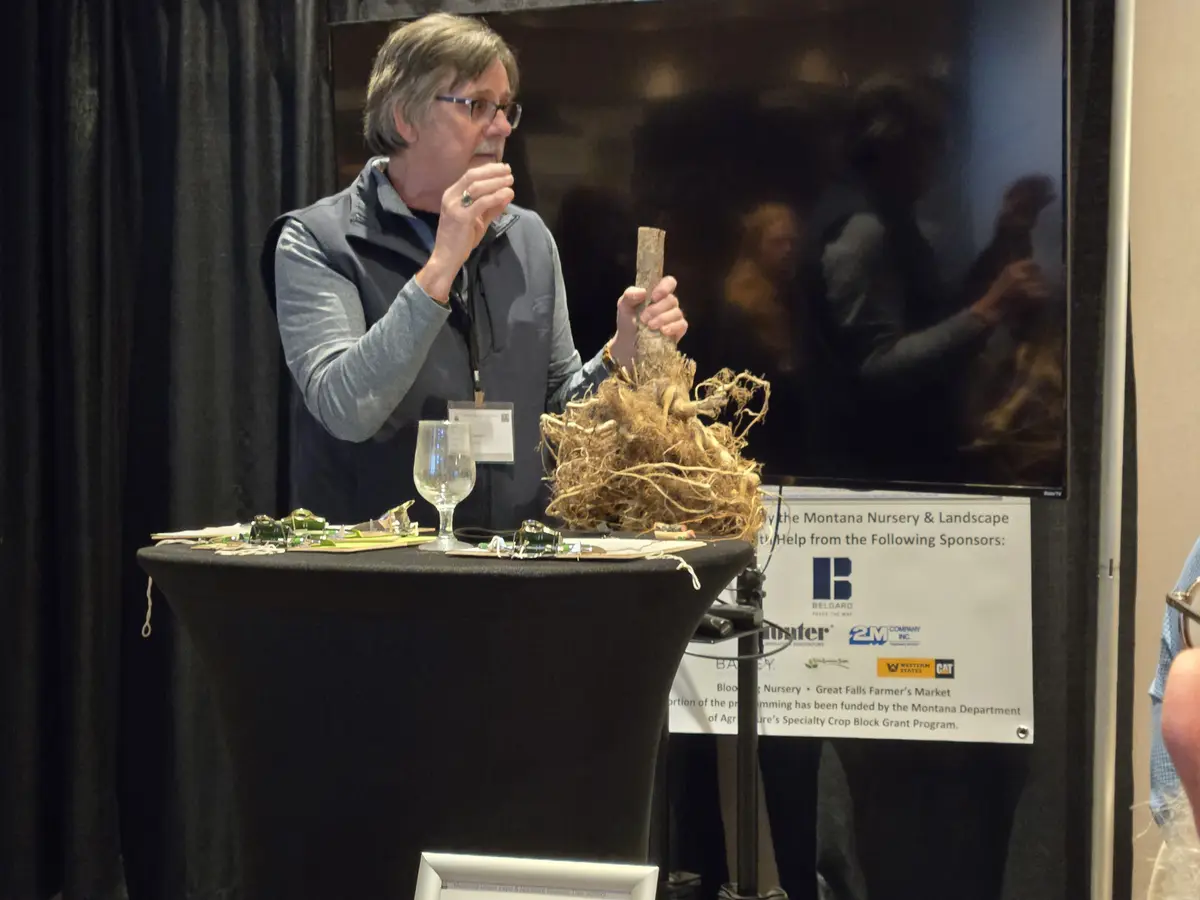 A man with glasses and a mustache stands behind a black table at what appears to be a conference or expo, holding a large…