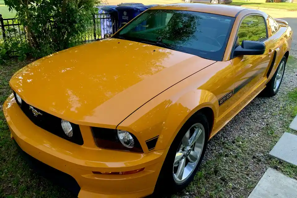 A bright yellow Ford Mustang GT/CS is parked on a gravel driveway next to grass and a sidewalk, with blue recycling bins…