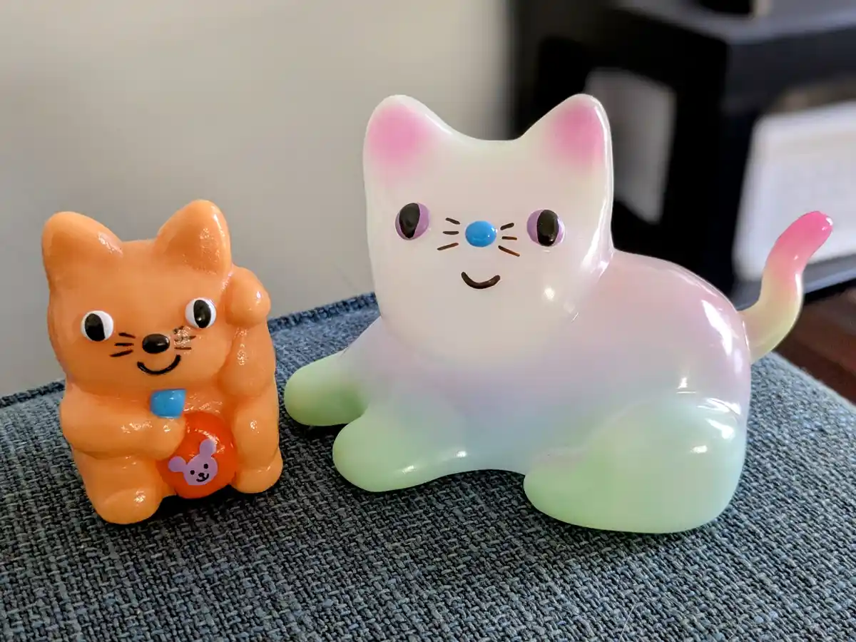 Two small, stylized cat figurines sit side by side on a textured gray-blue fabric. The left cat is solid orange with large…