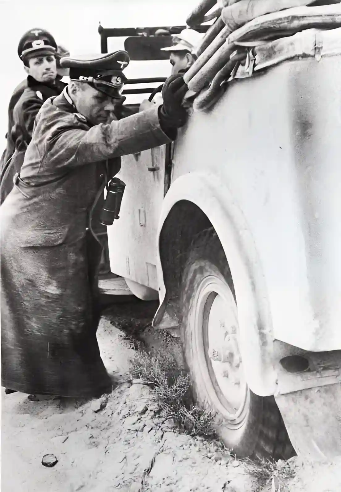 A black-and-white historical photo shows a man in a military uniform pushing a stuck military vehicle out of mud, while…