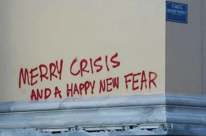 Red spray-painted text on a beige wall reads 'MERRY CRISIS AND A HAPPY NEW FEAR'. A blue street sign is visible in the upper…