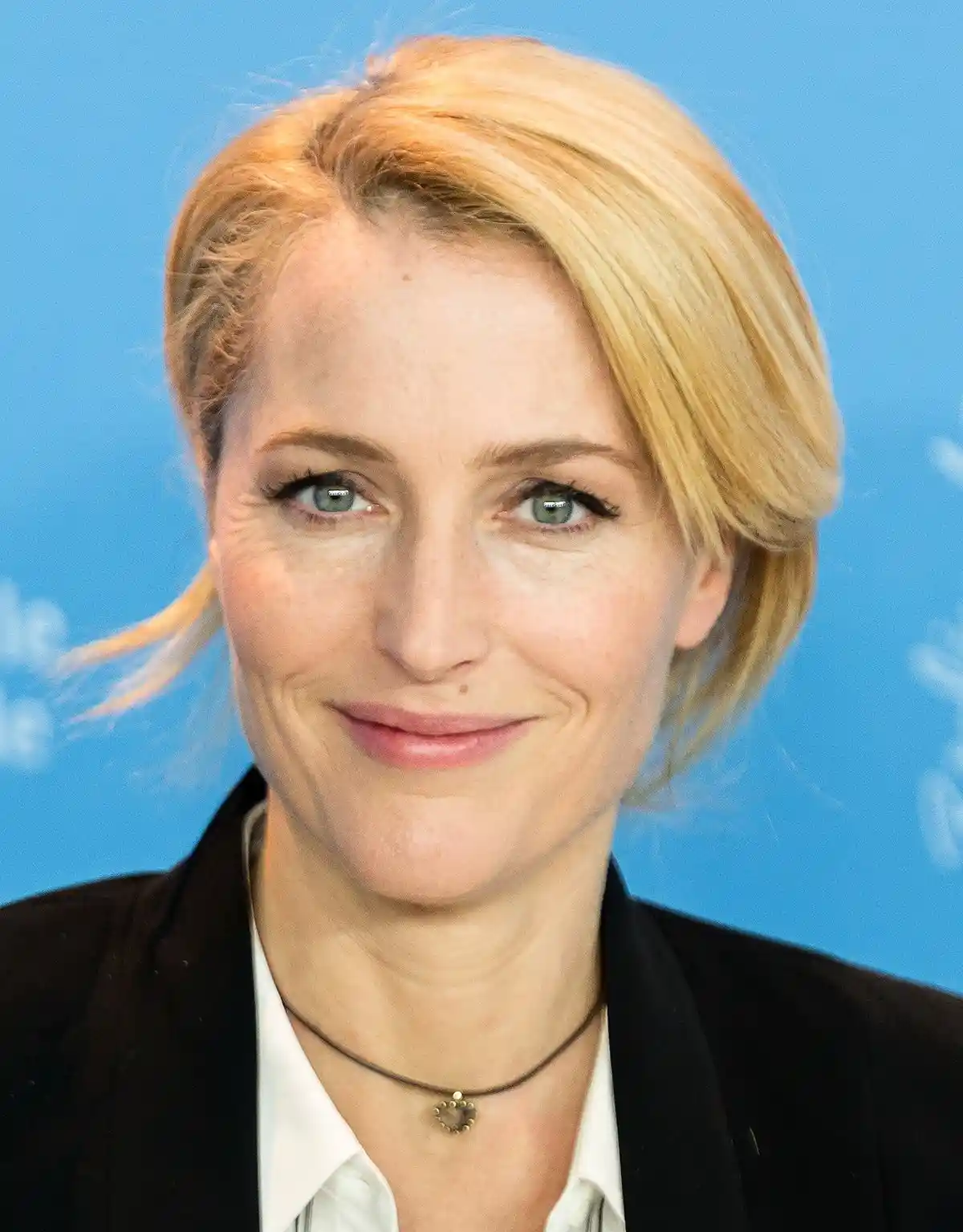 A close-up photo of a woman with blonde hair and green eyes, wearing a black blazer over a white shirt and a heart-shaped…