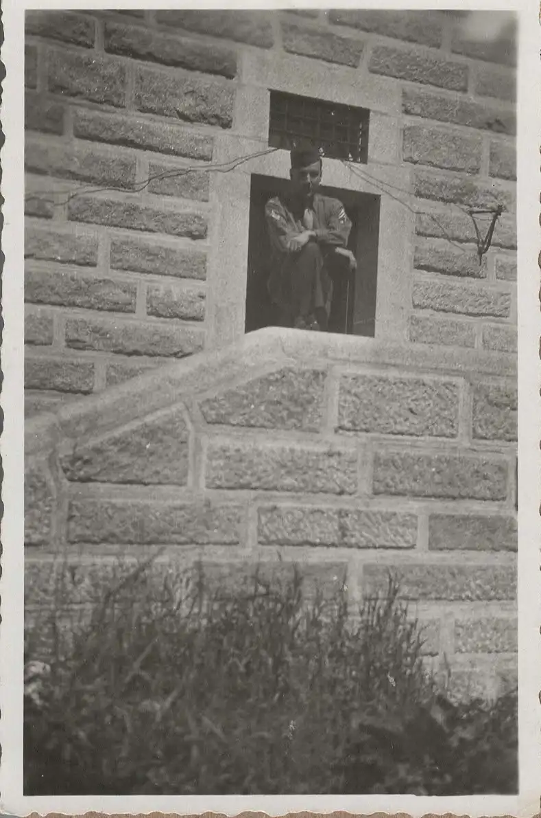 A black-and-white photo shows a man in a military uniform sitting in a small, barred window of a stone building, looking out…