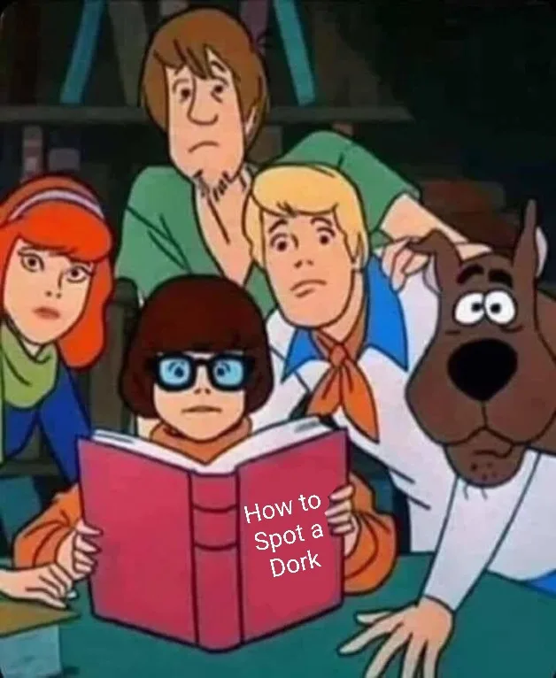 A still from the Scooby-Doo cartoon shows the main characters gathered around a table. In the center, Velma, wearing her…