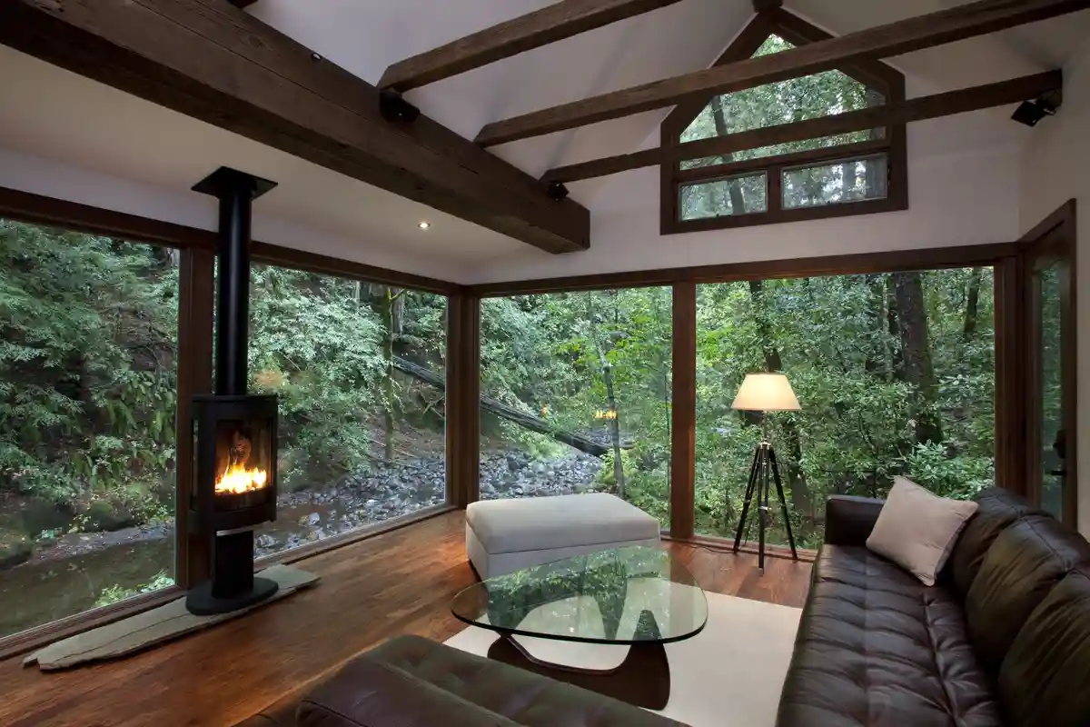 A cozy, modern living room with large windows overlooking a lush forest and a stream, featuring a lit wood-burning stove…