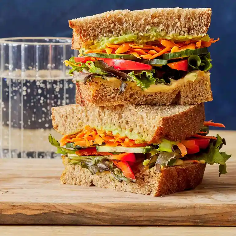 A visually appealing, stacked sandwich with layers of avocado, shredded carrots, cucumber, tomato, and mixed greens on whole…