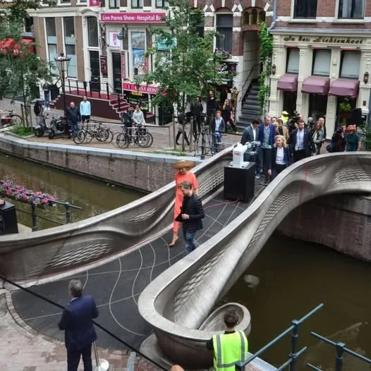 A photo of a modern, sculptural bridge over a canal in Amsterdam, with people walking across it. A woman in a bright orange…