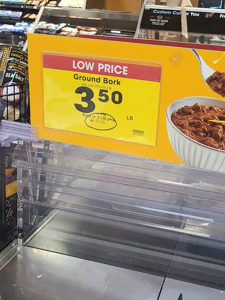 A supermarket meat aisle shelf with an empty transparent display case. A bright yellow price sign advertises 'Ground Bork'…