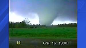 A grainy, low-resolution video still from a news broadcast shows a large, dark tornado touching down in a green field under…