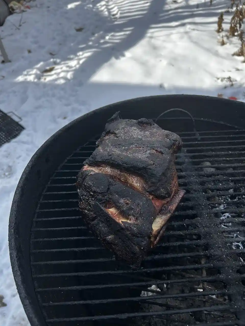 A large, heavily charred piece of meat sits on a black charcoal grill, surrounded by snow on the ground. The meat is…