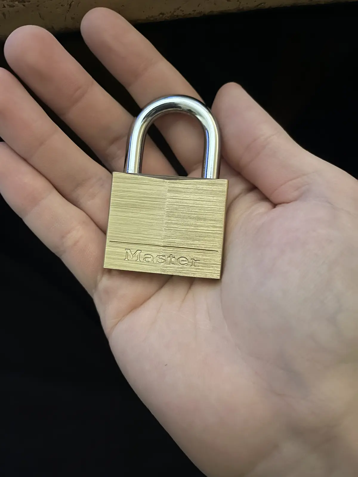 A close-up photo shows a person's open palm holding a small, brass-colored Master brand padlock with a shiny silver shackle…